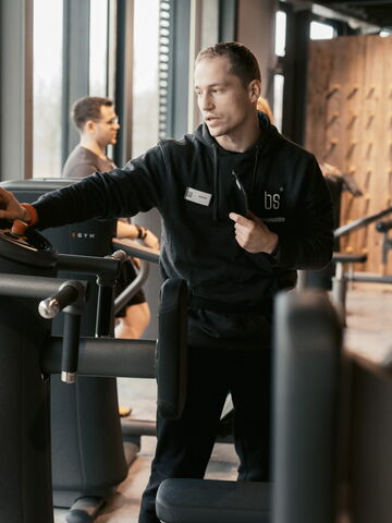 Ein Fitnessinstruktor erklärt einem Besucher die Bedienung eines modernen Trainingsgeräts im Fitnessstudio.