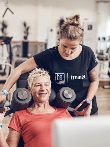 Eine Trainerin assistiert einer älteren Frau beim Krafttraining in einem modernen Fitnessstudio.