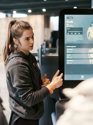 Eine Trainerin erklärt an einem Bildschirm die Kraftmessung und Muskelgruppen zu interessierten Zuhörern im Fitnessstudio.