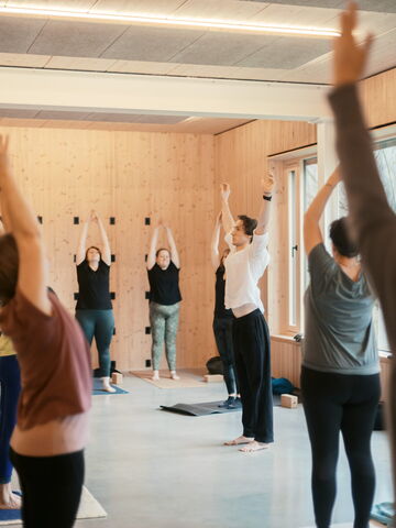 Eine Gruppe von Menschen praktiziert Yoga in einem hellen Raum, während sie die Arme zum Himmel strecken.