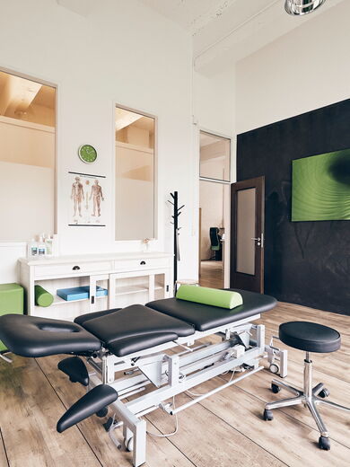 Ein helle Physiotherapiepraxis mit einer Liege, modernen Möbeln und grünen Akzenten an den Wänden.