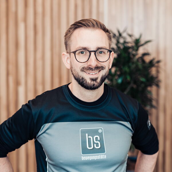 Ein lächelnder Mann in einem sportlichen Oberteil der Bewegungsstätte steht vor einer Holzpaneelwand und einer Pflanze.