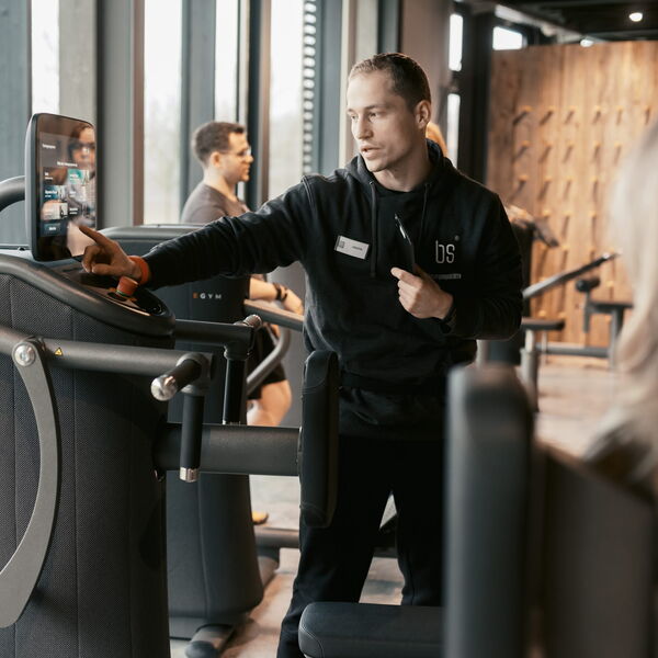 Ein Fitnessinstruktor erklärt einem Besucher die Bedienung eines modernen Trainingsgeräts im Fitnessstudio.
